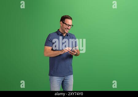 Hübscher junger Mann in Brille lächelt und liest Nachrichten über Handy auf grünem Hintergrund Stockfoto