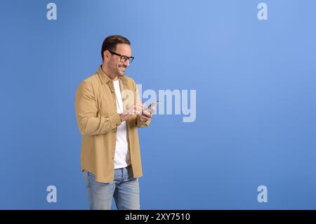 Glücklicher junger Geschäftsmann in Brille lächelt und schreibt online über Smartphone auf blauem Hintergrund Stockfoto