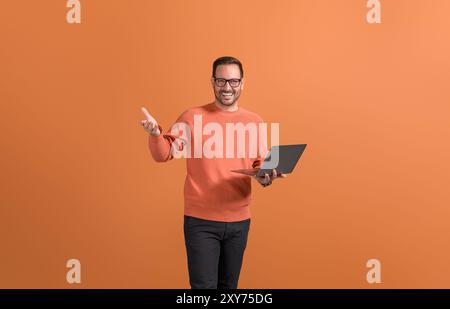 Hübscher Unternehmer mit Laptop und selbstbewusster Gesten, während er auf orangefarbenem Hintergrund steht Stockfoto