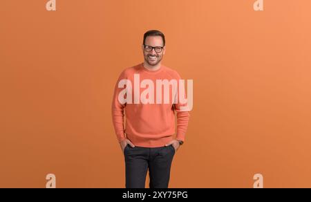 Porträt eines lächelnden männlichen Freelancers mit Händen in Taschen, die selbstbewusst auf orangefarbenem Hintergrund stehen Stockfoto