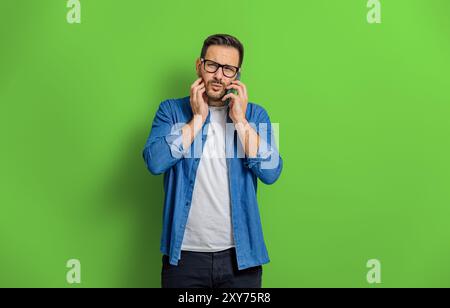Porträt eines jungen Mannes mit verwirrtem Ausdruck, der über Mobiltelefon vor grünem Hintergrund spricht Stockfoto