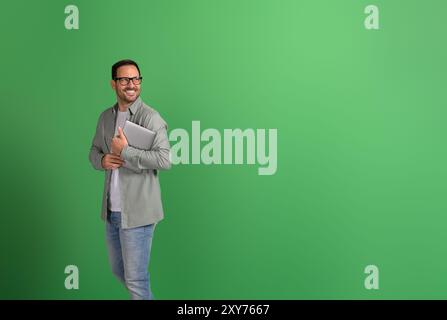 Positiver Geschäftsmann, der den Laptop hält und nachdenklich wegschaut, während er auf grünem Hintergrund steht Stockfoto
