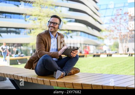 Lächelnder Profi mit Kopfhörern und einem digitalen Tablet, der wegblickt und in der Stadt auf der Bank sitzt Stockfoto