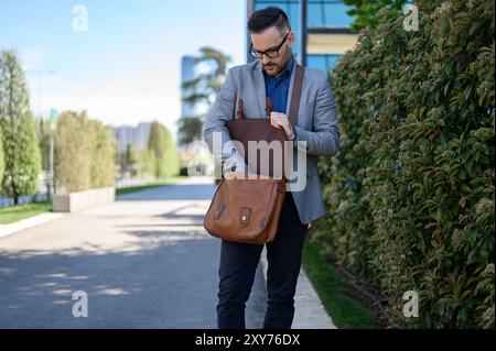 Junger Geschäftsmann in Brille, der braune Laptoptasche durchsucht, während er an Pflanzen auf der Straße der Stadt steht Stockfoto