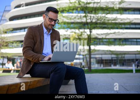 Konzentrierter Geschäftsmann, der Finanzberichte über Laptop analysiert, während er in der modernen Stadt auf der Bank sitzt Stockfoto
