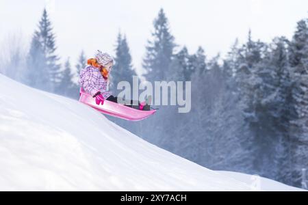 Ein kleines Mädchen, das auf einem rosafarbenen Schlitten den Berg hinunter rollt, die Geschwindigkeit wird klar. Pinke Jacke und farbenfroher Schal. Kleines Mädchen, das den Berg bergab rodelt Stockfoto