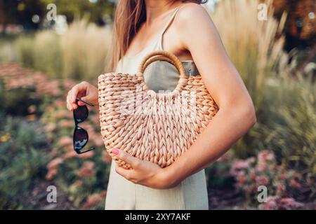Elegante, modische Frau, die im Sommer ein Kleid aus Leinen trägt und im Park eine Brieftasche aus Korb hält. Stilvolle Handtasche aus Bio-Anbau Stockfoto