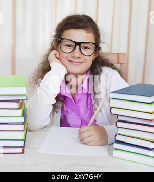 Das kleine brünette lächelnde Mädchen trägt eine Brille und macht Hausaufgaben Stockfoto