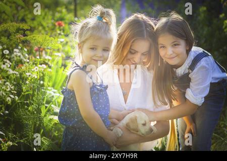 Glückliche Frau in weißer Bluse und ihre Töchter mit Welpen Labrador sitzen auf den Knien im Sommergarten Stockfoto