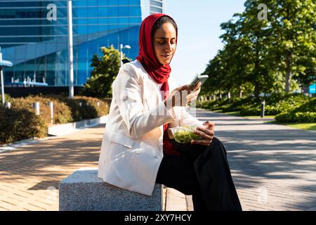 Schöne arabische Geschäftsfrau mit Hijab und elegantem Business-Anzug Porträt in der Stadt - erwachsene Frau aus dem Nahen Osten, die Salatschüssel zum Mitnehmen isst Stockfoto
