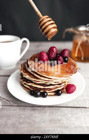 Saftige Pfannkuchen mit Beeren und Honig auf einem weißen Teller, Löffel, Glas, Holztisch, Kaffeetasse. Stockfoto