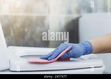 Zugeschnittene Bildansicht mit Nahaufnahme der Hände einer Frau mit Gummischutzhandschuhen. Hausfrau putzt Toilettenschüssel, Sitz mit pinkfarbenem Tuch, Wischtuch in der Badewanne Stockfoto