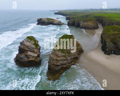 Gigantische, zerklüftete Felsen, die aus dem Meer ragen und von schäumendem Wasser umhüllt werden, aus der Luft, Praia das Catedrais, Playa de las Catedrales, als Catedrais bea Stockfoto