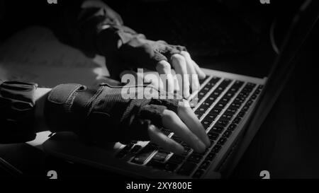 Abgeschnittene Aufnahme der Handschrift eines jungen Mannes auf dem Laptop während der Arbeit in einem schwarzen Bürozimmer. Hacker hacken den Server im Dunkeln Stockfoto