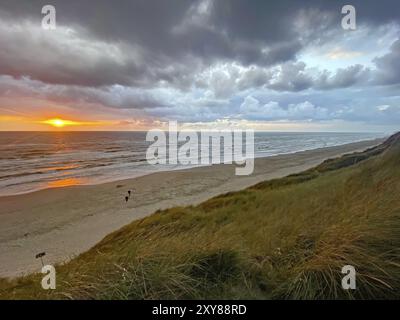 Sonnenuntergang über dem Meer unter einem dramatischen, bewölkten Himmel, mit Menschen am Strand und Grasdünen, Urlaub, Lokken, Jütland, Dänemark, Europa Stockfoto