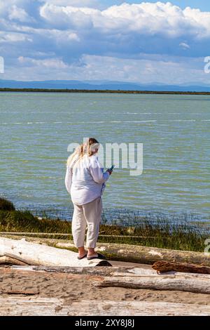 Eine junge Frau, die ihr Handy auf Treibholz in Steveston, Kanada, ansieht Stockfoto