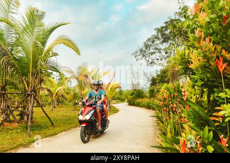 Glückliches lächelndes Paar Reisende, die während eines tropischen Urlaubs unter Palmen auf der Insel Phu Quoc, Vietnam, mit einem Motorradroller in Schutzhelmen fahren Stockfoto