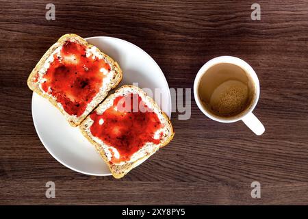 Blick von oben auf einem Teller mit Frühstück, zwei Brotscheiben mit Fruchtmarmelade, heißer Kaffee auf einem dunklen Holztisch Stockfoto