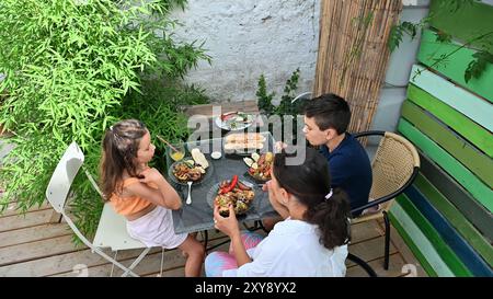 Eine Familie sitzt um einen Tisch auf einer Terrasse und genießt eine gemeinsame Mahlzeit inmitten von lebhaftem Grün. Eine Szene der Wärme und Zweisamkeit. Stockfoto