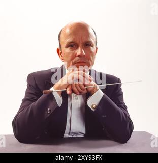 Ein seriöser Dirigent mittleren Alters in formeller Kleidung legt Hände auf den Stab und blickt nachdenklich auf das Studio-Porträt mit dunklem Hintergrund und dramatischer Beleuchtung Stockfoto