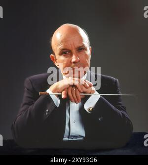 Ein seriöser Dirigent mittleren Alters in formeller Kleidung legt Hände auf den Stab und blickt nachdenklich auf das Studio-Porträt mit dunklem Hintergrund und dramatischer Beleuchtung Stockfoto