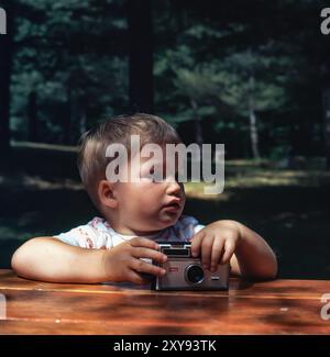 Neugieriger Kleinkind mit einer alten Instamatic-Kamera, der draußen an einem hölzernen Picknicktisch sitzt und in einem sonnendurchfluteten Wald beiseite blickt. Ruhig! Stockfoto