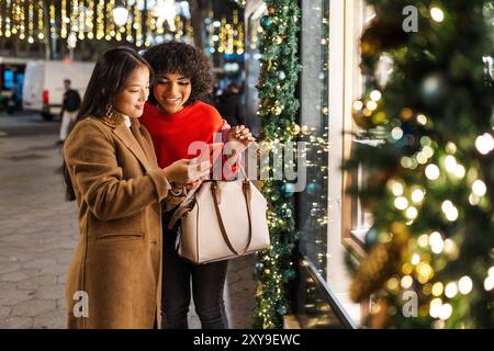 Zwei junge Frauen, die ihr Smartphone benutzen, in der weihnachtlich geschmückten Stadt in der Nacht Stockfoto