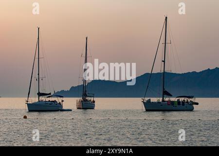 Segelboote auf orangefarbenem Hintergrund bei Sonnenuntergang. Segelboote auf dem ruhigen Mittelmeer. Segelboote vor Anker im Golf von Porto auf Korsika. Stockfoto