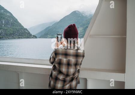 Frau genießt Fjordlandschaften beim Fotografieren mit Smartphone auf der norwegischen Fähre mit nebelgrünen Bergen im Hintergrund. Szene fängt das Wesen von Re ein Stockfoto