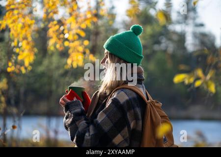 Eine glückliche träumende Frau wärmt draußen im kalten Spätherbst die Hände an einer Thermoskanne und schlendert durch den Wald. Stockfoto