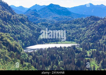 Toller See und Wald in Bayern, Deutschland. Ruhiger See, umgeben von üppigen Bäumen und majestätischen Bergen unter einem klaren blauen Himmel. Der See ist von Tre umgeben Stockfoto