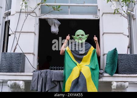 Eine Person, die eine Maske trägt und in die jamaikanische Flagge gehüllt ist, beobachtet die Notting Hill Carnival Parade vorbei. Stockfoto