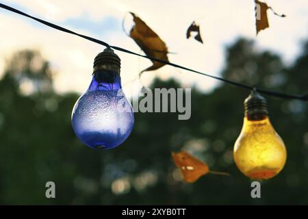 Eine Kette von Lichtern hängt am Baum. Gartenparty. Romantischer Ort. Bunte Glühbirnen. Abendstimmung, festliche Stimmung. Wochenende, Urlaub oder jeden Stockfoto