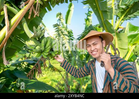 Ein indonesischer Bauer hält Bananen und lächelt. Er trägt einen Strohhut und ein gestreiftes Hemd Stockfoto