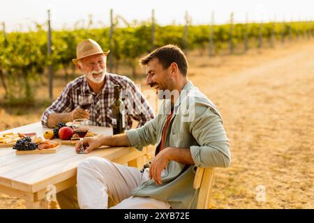 Zwei Freunde genießen die reichhaltigen Aromen von lokalen Weinen und köstlichen Snacks an einem rustikalen Tisch im Freien, während sie sich bei Sonnenuntergang im goldenen Glanz des Weinbergs erfreuen Stockfoto