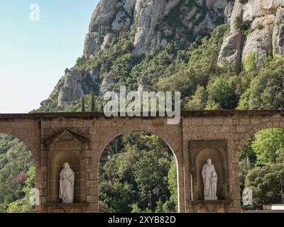 Steinstatuen unter Bögen vor einer felsigen, bewaldeten Berglandschaft und klarem Himmel, montserrat, spanien Stockfoto