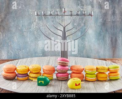 Eine Hanukkah-Menora mit bunten Macarons und Dreidels auf einer Holzoberfläche, die die festliche Jahreszeit feiert. Stockfoto