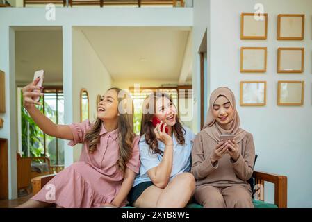 Drei indonesische Frauen sitzen auf einer Couch und zwei von ihnen Videoanrufe auf ihren Telefonen. Die anderen Frauen reden über ihre Telefone. Die Szene ist li Stockfoto