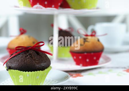 Liebevoll dekorierte Muffins auf einem Couchtisch mit gestickter Tischdecke. Im Vordergrund ein Schokoladenmuffin mit roter Schleife und grüner Hülle. Ein et Stockfoto