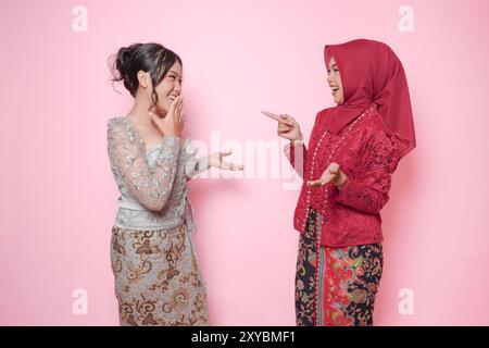 Zwei asiatische Frauen als enge Freundinnen, die moderne Kebaya tragen und ein lustiges Gespräch führen, isoliert über rosa Hintergrund. Stockfoto