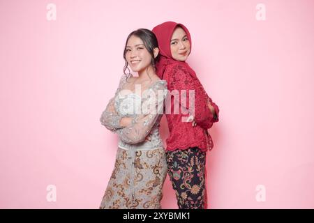 Zwei lächelnde asiatische Frauen im modernen Kebaya-Kleid posieren und sich auf den Rücken lehnen, isoliert durch rosa Hintergrund. Stockfoto