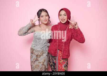 Zwei fröhliche junge asiatische Frau, die modernes Kebaya trägt, mit Friedenszeichen, isoliert auf rosa Hintergrund. Stockfoto