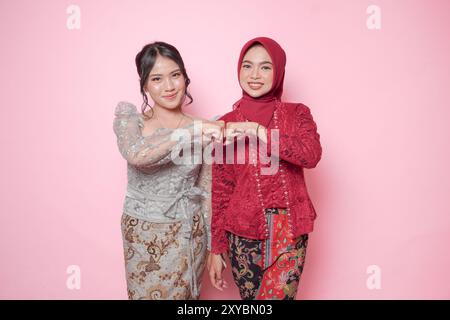 Lächelnde asiatische junge Frauen, die moderne Kebaya tragen, zeigen ein Faustzeichen, isoliert auf rosa Hintergrund. Stockfoto