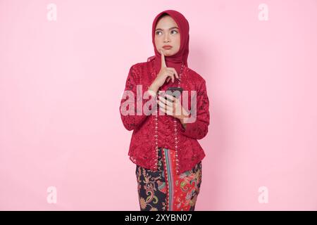 Nachdenkliche junge indonesische Frau mit modernem Kebaya und Hijab, die ihr Smartphone hält, während sie über isolierten rosa Hintergrund nachdenkt. Stockfoto