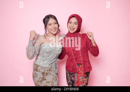 Zwei aufgeregte asiatische Frauen tragen ein modernes Kebaya-Kleid mit einem glücklichen, erfolgreichen Ausdruck, isoliert durch rosa Hintergrund. Stockfoto