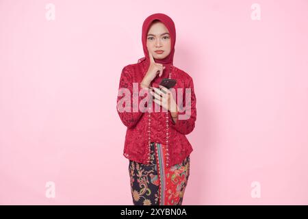 Nachdenkliche junge indonesische Frau mit modernem Kebaya und Hijab, die ihr Smartphone hält, während sie über isolierten rosa Hintergrund nachdenkt. Stockfoto