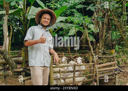 Glücklicher asiatischer Bauer, der mit der Hand nach oben gezeigte Zustimmungsgeste mit Viehziegen für Opfer dahinter zeigt. EID Al Adha Konzept. Stockfoto