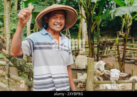 Ein glücklicher indonesischer Viehzüchter, der die Daumen hochgibt, mit Viehziegen für Opfer hinter ihm. EID Al Adha Konzept. Stockfoto