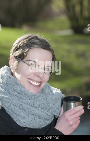 Lächelnde Frau in einem warmen Winterschal, die einen warmen Becher mit heißem Getränk in den Händen hält, während sie draußen in einem Park sitzt. Lächelnde Frau in einem warmen Winter Stockfoto