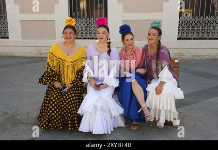 Vier Frauen in bunten Flamenco-Kleidern und Blumen im Haar sitzen auf einer Bank auf einer Straße in traditioneller spanischer Mode, Feria de la Manzanilla, Stockfoto